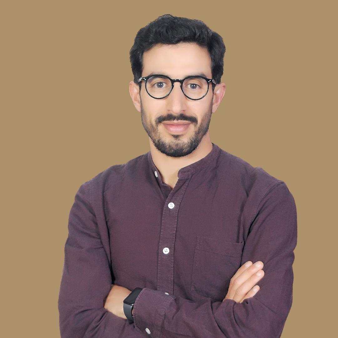 Portrait of a man with dark hair and glasses, arms crossed, wearing a maroon button-down shirt against a solid brown background.