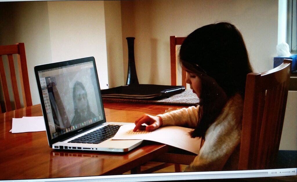 Girl at a wooden table typing on a silver laptop while writing on a sheet of paper, in a dining room setting with chairs and a window in the background